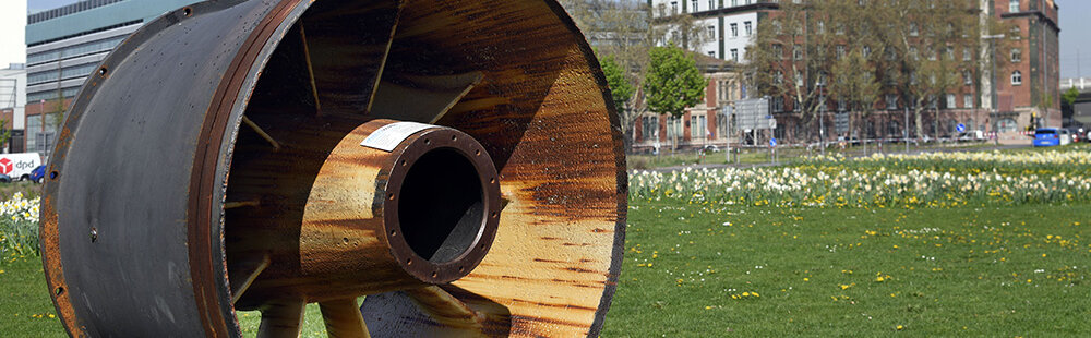 In einem städtischen Park steht ein massives, metallisches Turbinenrad, das an eine industrielle Vergangenheit erinnert.