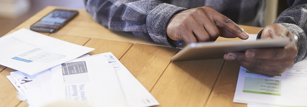 Auf dem Tisch liegen mehrere Briefe, während eine Hand ein Tablet bedient und ein Smartphone im Hintergrund sichtbar ist.