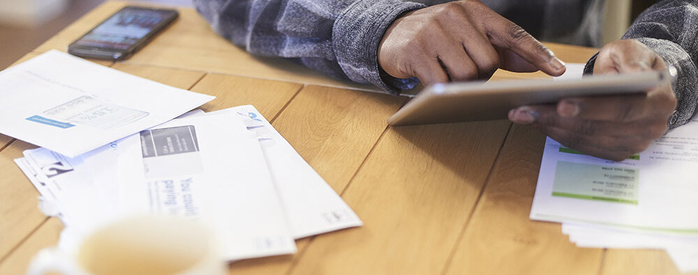 Auf dem Tisch liegen mehrere Briefe, während eine Hand ein Tablet bedient und ein Smartphone im Hintergrund sichtbar ist.