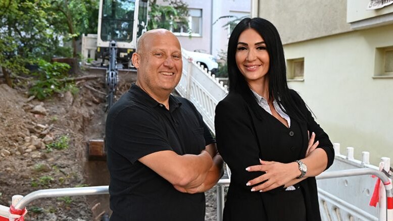 Zwei Personen posieren fröhlich vor einer Baustelle, während ein Bagger im Hintergrund auf einen Graben hindeutet.