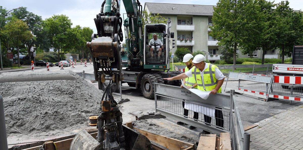 Zwei Bauarbeiter in Sicherheitswesten besprechen Pläne am Baustellenrand, während ein Bagger im Hintergrund bereitsteht.