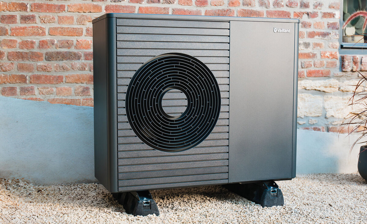 Eine moderne Wärmepumpe steht auf Kiesboden vor einer Backsteinmauer.