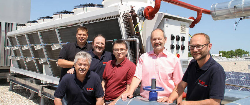 Sechs Personen stehen lächelnd vor einer großen technischen Anlage auf einem Dach, umgeben von Sonnenkollektoren.