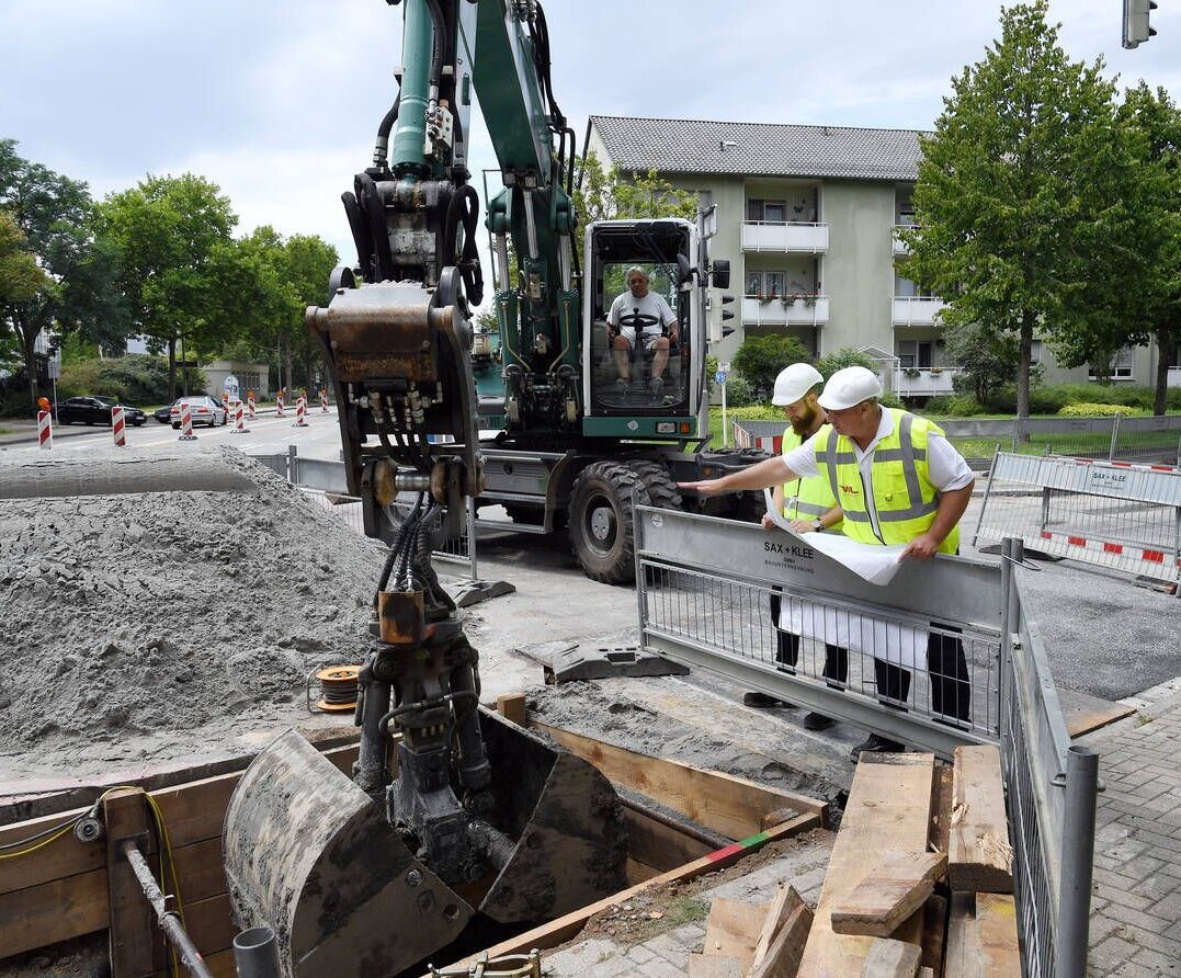 Zwei Bauarbeiter in Sicherheitswesten besprechen Pläne vor einer Baustelle mit einem Bagger im Hintergrund.