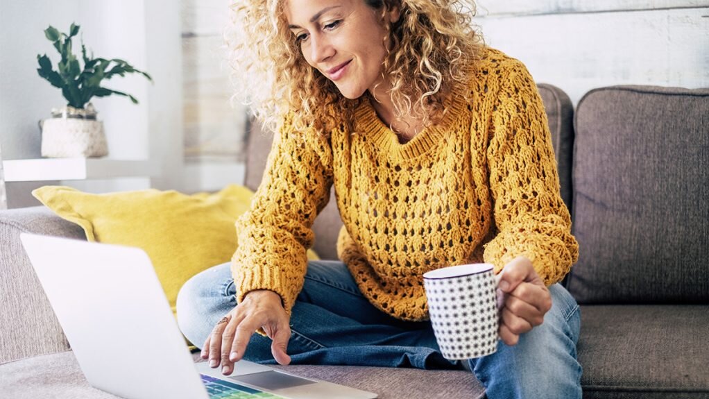 Die Frau in einem gestrickten Pullover sitzt entspannt auf dem Sofa, während sie auf den Bildschirm ihres Laptops schaut und Kaffee trinkt.
