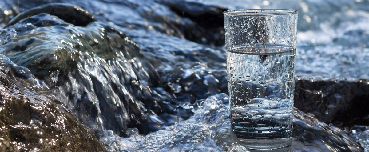 Ein transparentes Glas mit Wasser steht inmitten von fließendem Wasser, das die Steine umspült und eine lebendige Atmosphäre schafft.