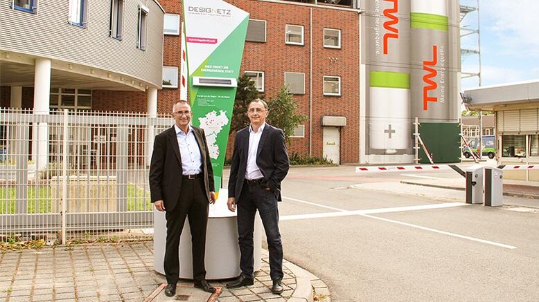 Die Szene zeigt zwei Herren in Anzügen, die lächelnd vor einem Firmengebäude und einem auffälligen Werbebanner posieren.