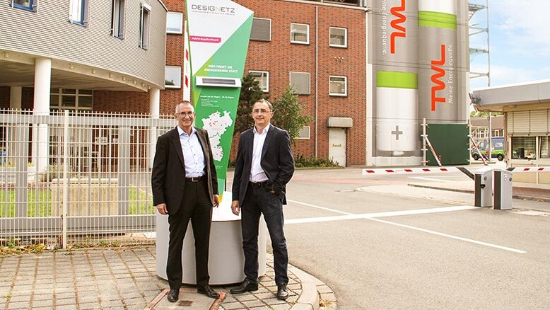 Die Szene zeigt zwei Herren in Anzügen, die lächelnd vor einem Firmengebäude und einem auffälligen Werbebanner posieren.