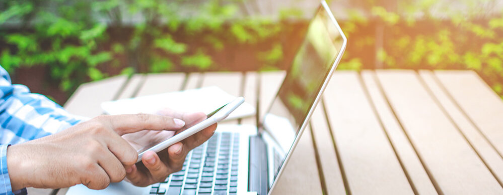 Eine Person sitzt an einem Tisch im Freien und nutzt ein Smartphone, während ein Laptop daneben steht.