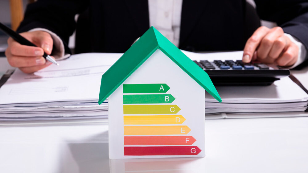 Ein Modellhaus mit einer Energieeffizienzskala von A bis G steht auf einem Tisch, während eine Person mit einem Stift Notizen macht.