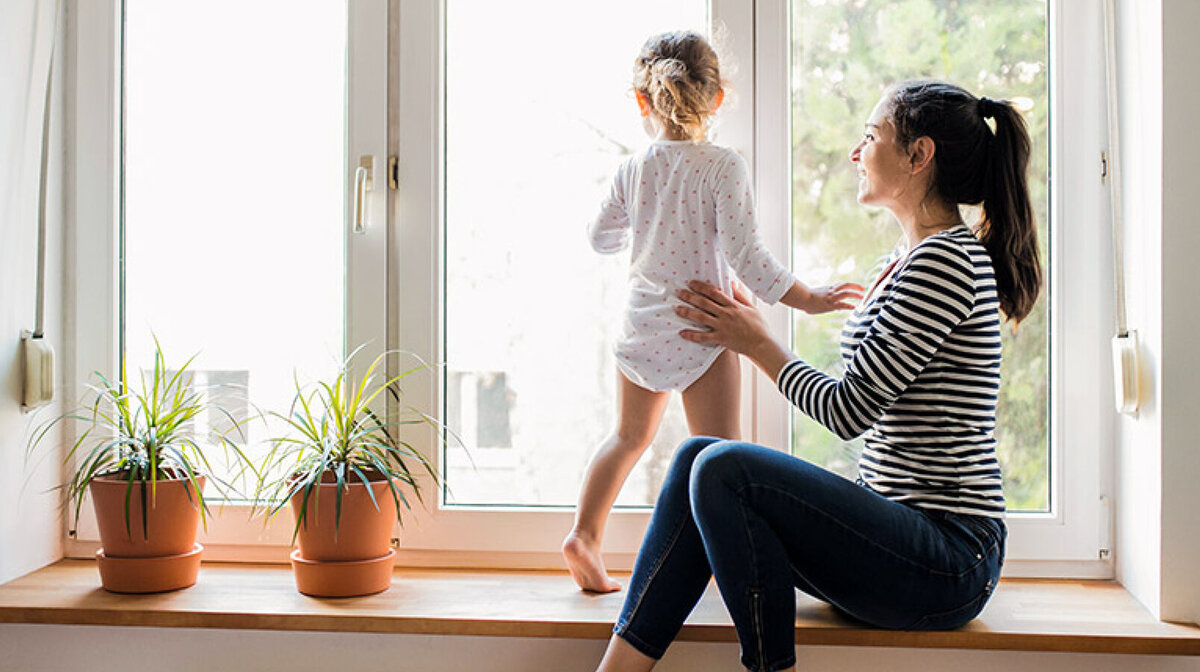 Ein kleines Mädchen steht auf der Fensterbank und blickt nach draußen, während ihre Mutter liebevoll neben ihr sitzt.