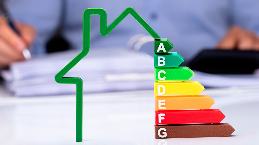 Die Energieeffizienzskala zeigt von grün bis braun, während im Hintergrund eine Person mit Unterlagen arbeitet.