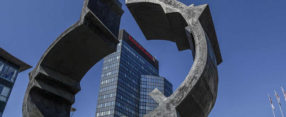 Eine große, moderne Skulptur aus Beton steht vor einem Hochhaus unter einem strahlend blauen Himmel.