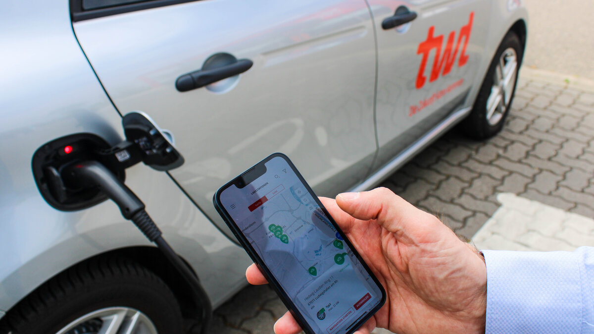 Hand hält Smartphone mit geöffneter App vor ein Elektroauto mit rotem Logo 'twl', das an einer Ladestation angeschlossen ist.