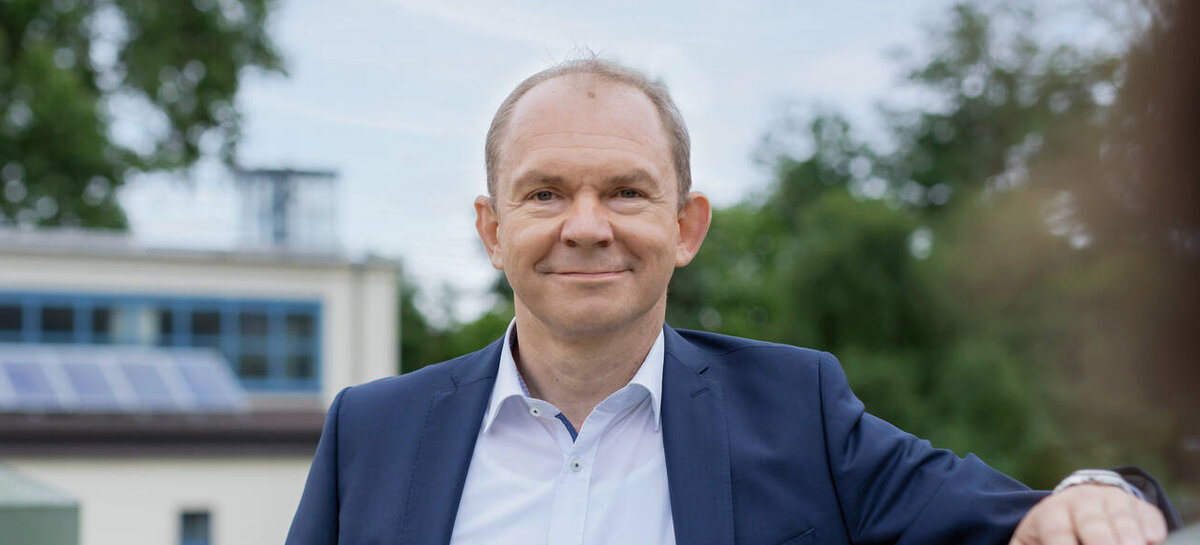 In einem Park steht ein lächelnder Mann in formeller Kleidung, während im Hintergrund Bäume und ein Gebäude sichtbar sind.