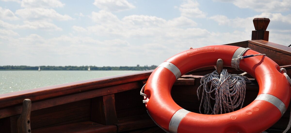 Ein orangefarbener Rettungsring liegt auf dem Holzdeck eines Bootes, während sanfte Wellen im Hintergrund glitzern.