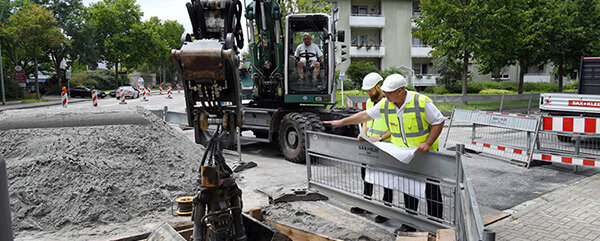 Zwei Bauarbeiter in Sicherheitskleidung besprechen Pläne, während ein Bagger in der Nähe eine Grube aushebt.