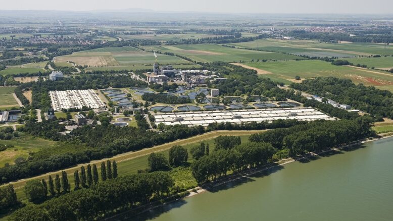 Eine Luftaufnahme zeigt eine weitläufige Landschaft mit Wasserbecken und grünen Feldern, umgeben von Bäumen und einem Fluss.