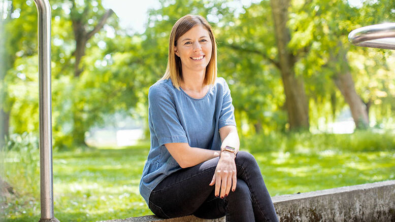 Eine lächelnde Frau sitzt entspannt auf einer Steinmauer in einem sonnigen Park, umgeben von grünen Bäumen.