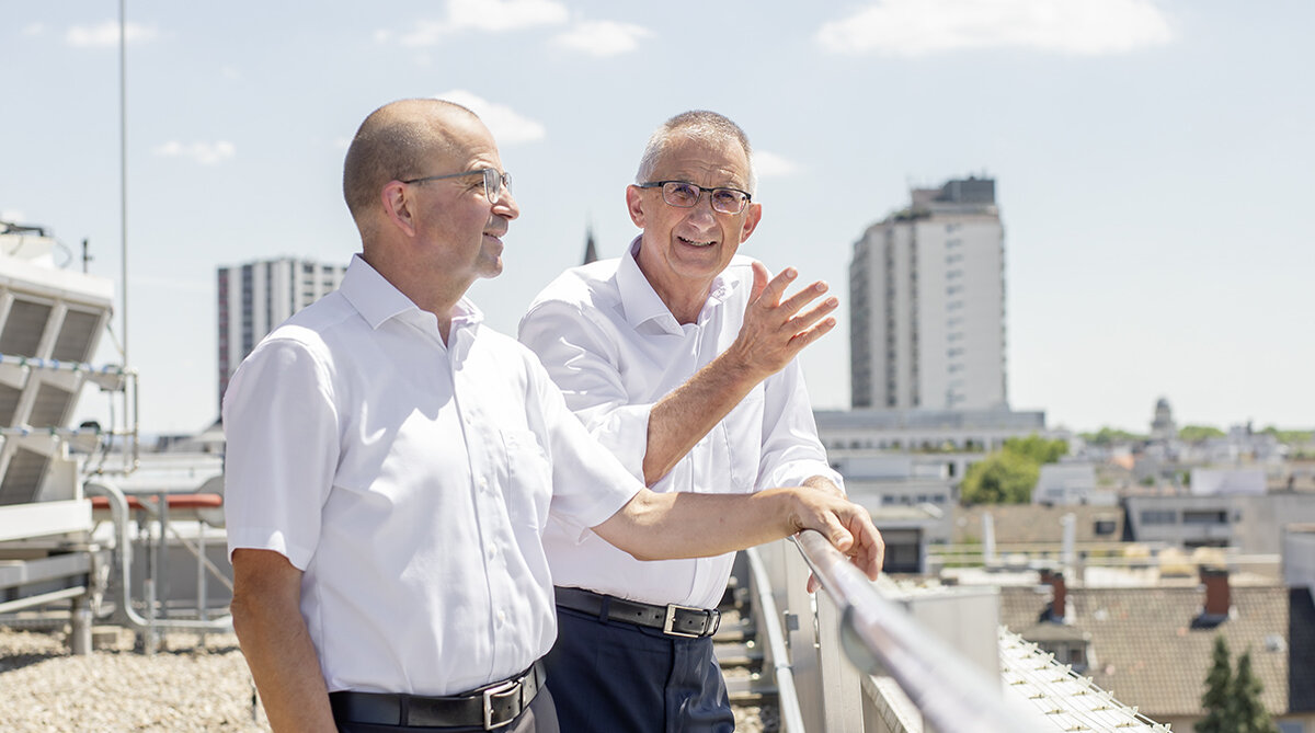 Die Szene zeigt zwei Herren in weißem Hemd auf einer Dachterrasse in einer Großstadt