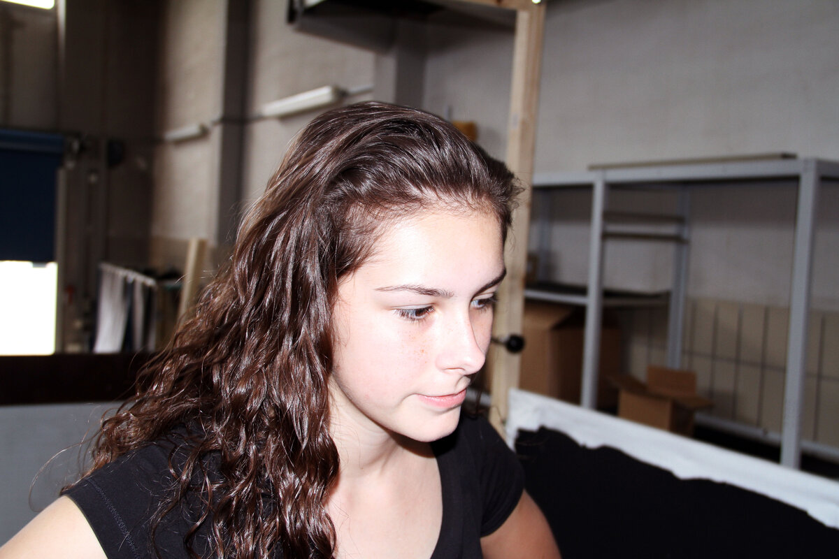 In einem industriellen Umfeld betrachtet ein Mädchen mit langen, lockigen Haaren aufmerksam etwas außerhalb des Bildes.