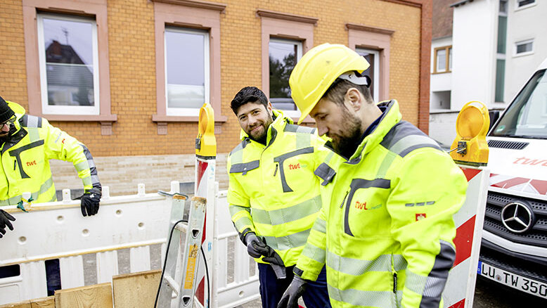 Drei Arbeiter in leuchtend gelben Sicherheitsjacken stehen an einer Baustelle und arbeiten an einem Absperrzaun.