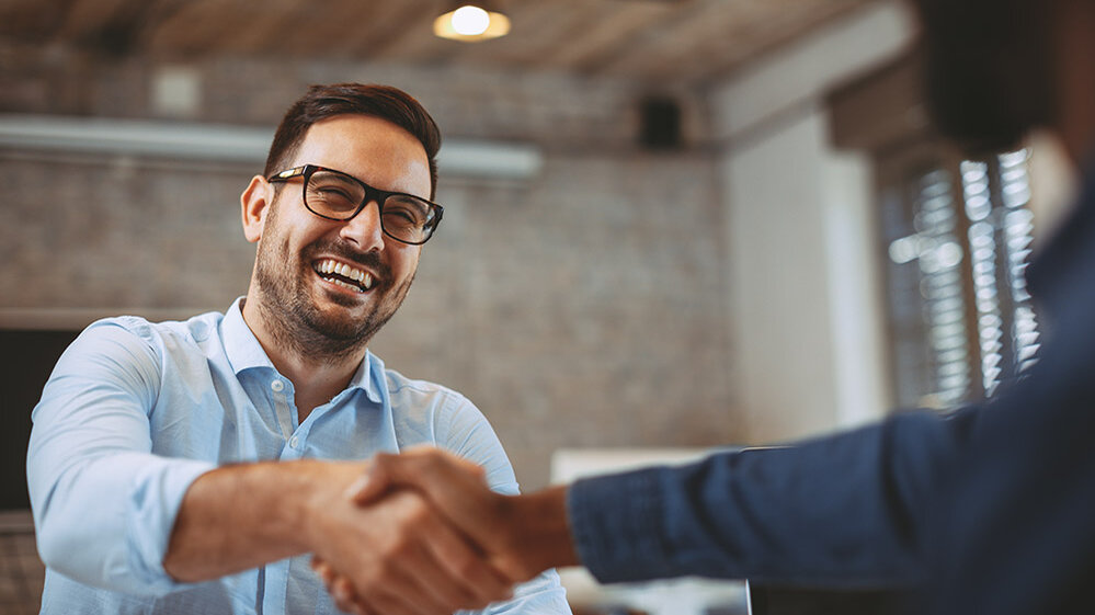 Ein lächelnder Mann in einem hellen Hemd schüttelt einem anderen Mann die Hand in einem modernen Büro.