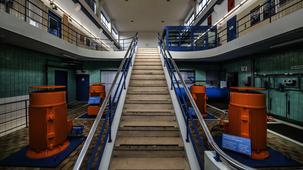 Die Szene zeigt eine industrielle Umgebung mit orangefarbenen Pumpen, einer Treppe und großen Fenstern, die Tageslicht hereinlassen.