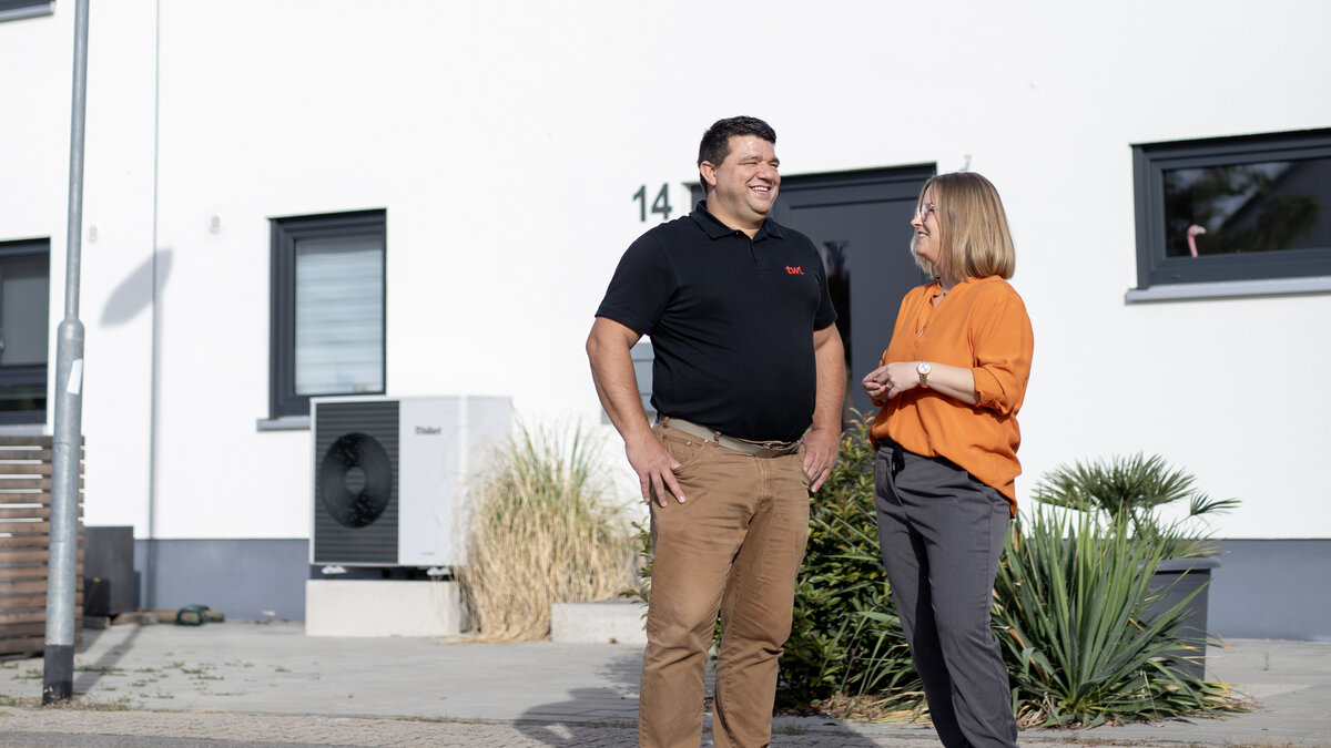 Ein Mann und eine Frau stehen lächelnd vor einem modernen, weißen Haus mit großen Fenstern und einer Wärmepumpe.