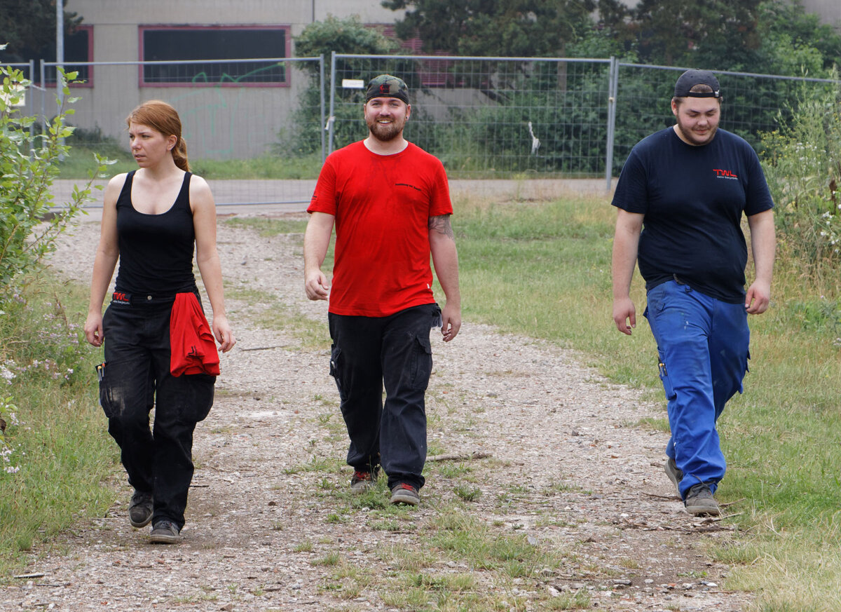 Drei Personen gehen auf einem Schotterweg, umgeben von grüner Vegetation und einem Baustellenzaun im Hintergrund.