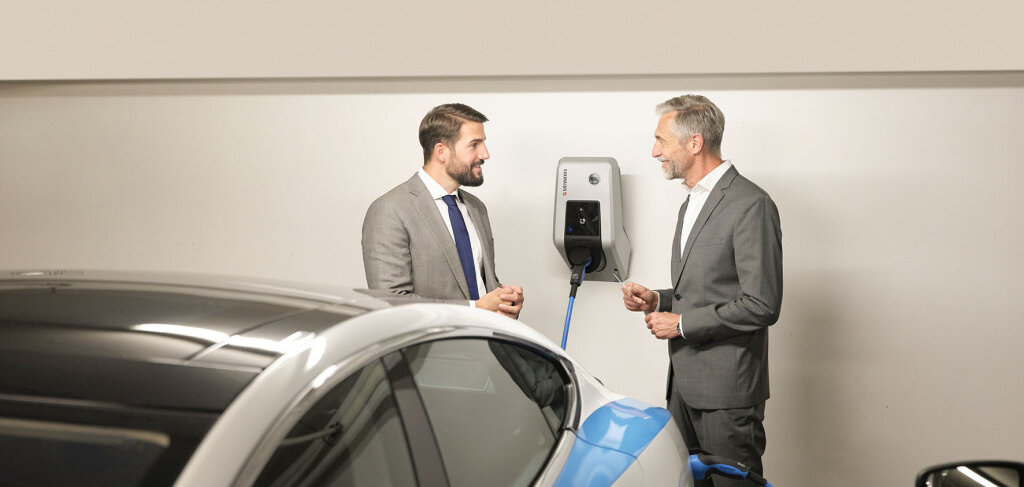 Zwei Männer in Anzügen unterhalten sich lächelnd neben einem Elektroauto, das an einer Ladestation angeschlossen ist.