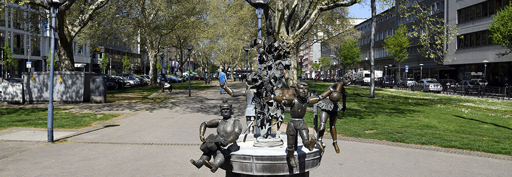 Im Park steht ein kunstvoller Brunnen mit einer Skulptur von Kindern, während die Sonne scheint.