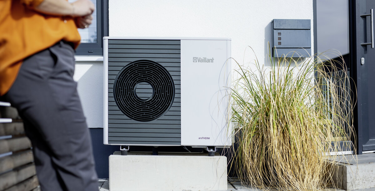 Im Vordergrund ist eine stilvolle Wärmepumpe zu sehen, während im Hintergrund eine Person entspannt an der Wand lehnt.