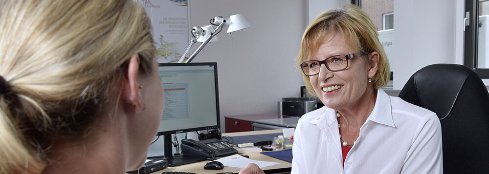 Eine blonde Frau sitzt an einem Schreibtisch und spricht mit einer Beraterin, die freundlich lächelt und einen Stift hält.