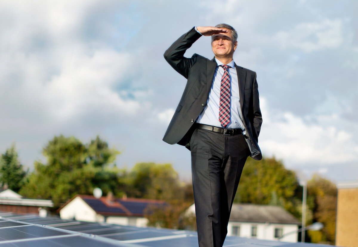 Auf einem Dach voller Solarpanels steht ein lächelnder Mann, der mit einer Hand seine Augen schützt und in die Landschaft blickt.