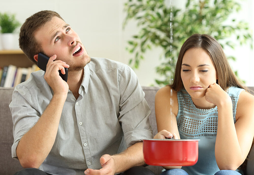Ein Mann telefoniert gestresst, während eine Frau frustriert auf einen Topf schaut, in den Wasser tropft.