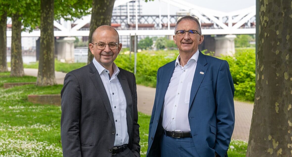 Zwei Männer in Anzügen stehen lächelnd auf einem grünen Weg, umgeben von Bäumen und einer Brücke im Hintergrund.