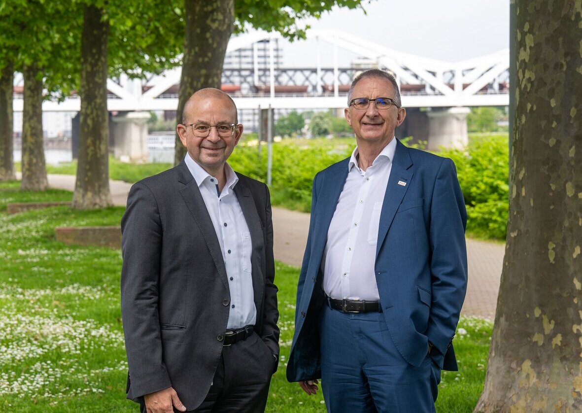 Zwei Männer in Anzügen stehen lächelnd auf einem grünen Weg, umgeben von Bäumen und einer Brücke im Hintergrund.