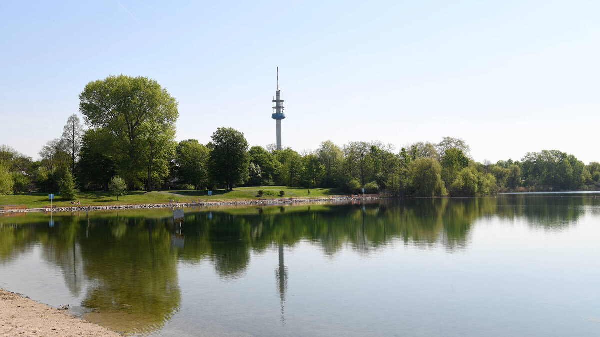 Ein ruhiger See spiegelt die umgebenden Bäume und den hohen Aussichtsturm an einem klaren, sonnigen Tag wider.