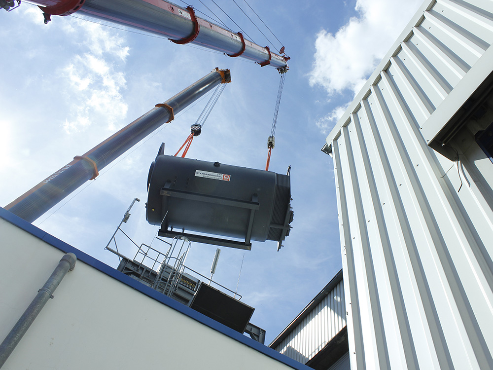 Ein grauer Behälter wird von einem Kran in die Höhe gezogen, mit einem modernen Industriegebäude im Hintergrund.