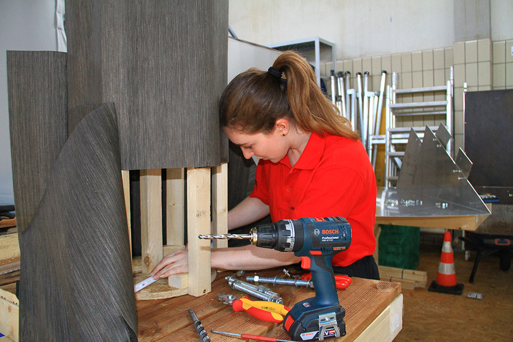 Eine junge Frau in einem roten Shirt arbeitet konzentriert an einem Holzprojekt, während sie ein Werkzeug benutzt.