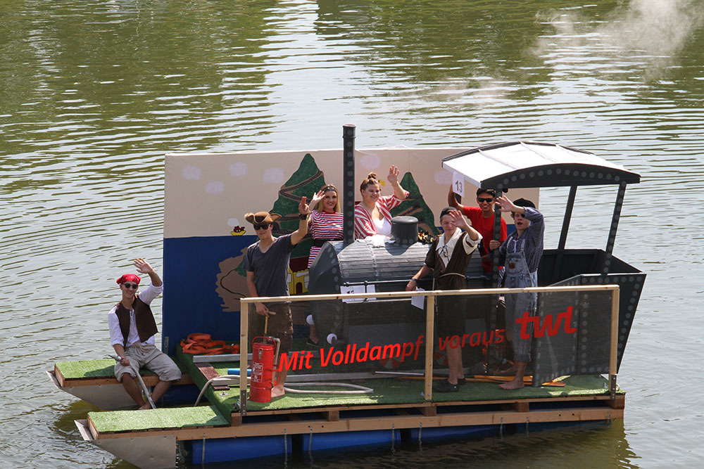Auf einem schwimmenden Boot posieren mehrere Personen in Kostümen, während sie in die Kamera winken und lachen.