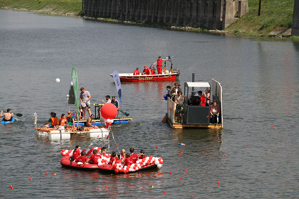 Ein Floß mit fröhlichen Menschen und bunten Dekorationen schwimmt auf dem Wasser, umgeben von weiteren Booten.