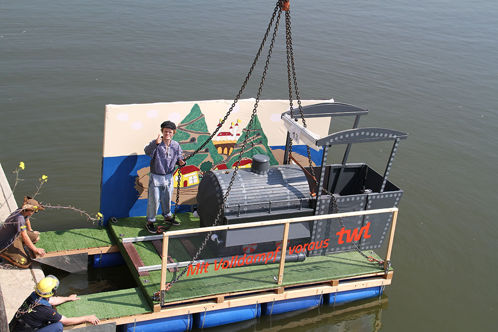 Ein Junge steht auf einer schwimmenden Plattform mit einem großen, grauen Dampfschiff und lächelt in die Kamera.