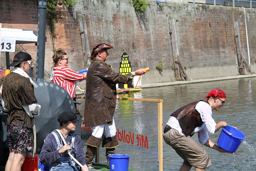 Die Szene zeigt mehrere verkleidete Personen, die an einem sonnigen Tag an einem Gewässer mit Eimern spielen.