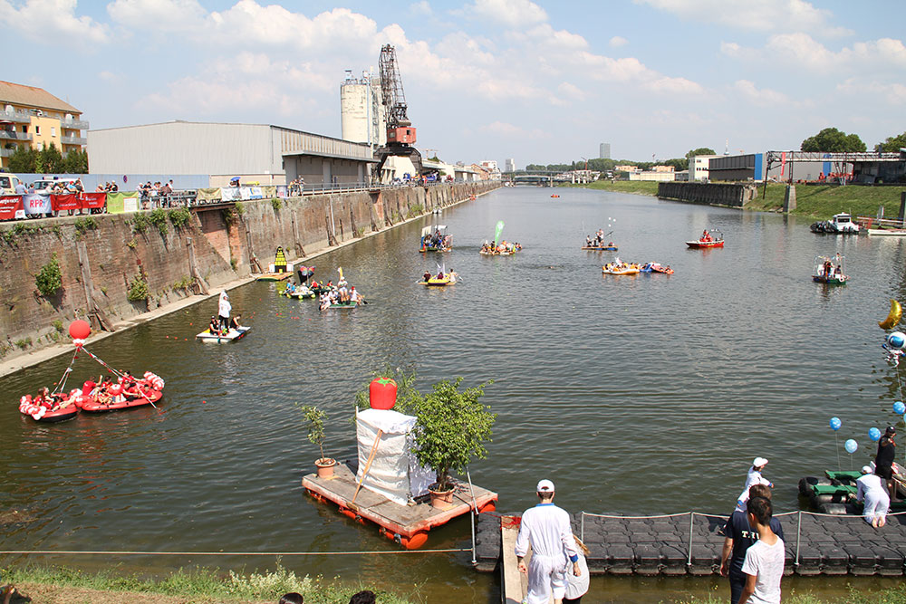 Eine lebhafte Szene am Wasser mit bunten Booten, die an einem sonnigen Tag an einem Kanal fahren, umgeben von Zuschauern.