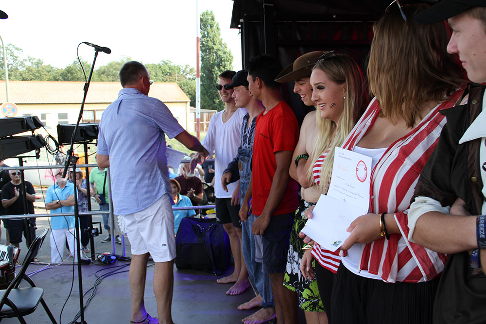 Ein Mann überreicht einer Gruppe von Jugendlichen auf einer Bühne Urkunden, während das Publikum im Hintergrund begeistert zuschaut.