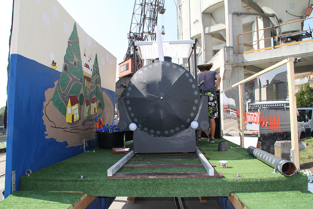 Eine bunte Kulisse zeigt eine fantasievolle Lokomotive auf einem Wagen, umgeben von einer malerischen Landschaft mit Hütten.