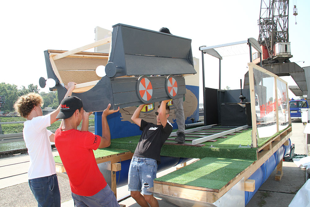 Eine Gruppe von Männern heben gemeinsam ein großes, graues Modell mit Rädern an, während sie auf einer Plattform stehen.