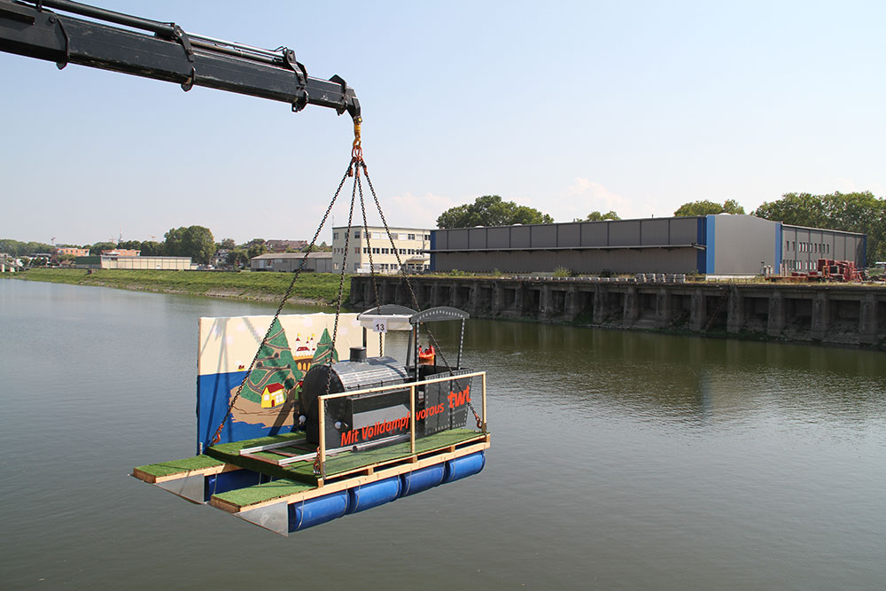 Ein Kran hebt eine schwimmende Plattform mit einem dekorierten Boot über einem ruhigen Fluss, umgeben von Gebäuden.